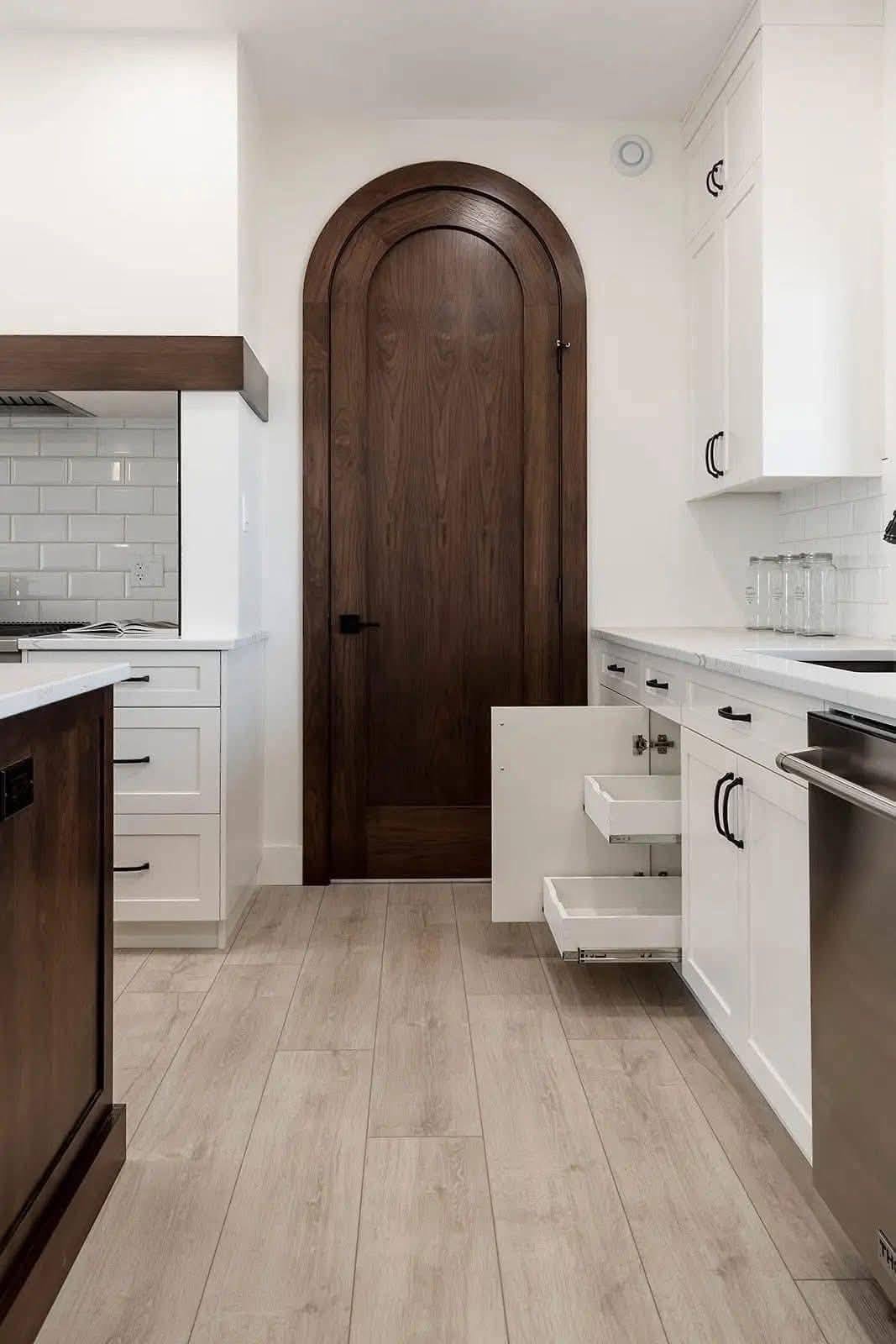 Sleek kitchen with white cabinets, subway tile backsplash, and light wood flooring. Notable arched wooden door adds warmth, open drawers suggest functionality.