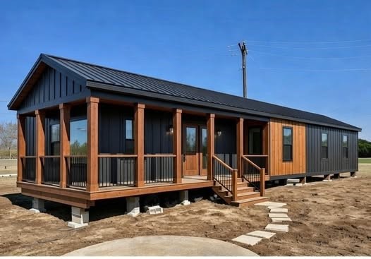 Modern tiny house with dark siding and warm wood accents, situated on a dirt lot. It features a porch with steps and a gable roof under a clear sky.