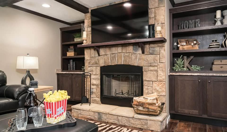A cozy living room features a stone fireplace with a large TV above, dark wood shelves with decor, and a black leather couch. Popcorn and glasses on a table add a relaxed, welcoming vibe.