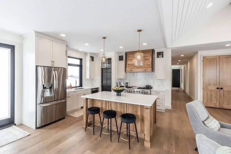 Modern kitchen with a large island and three black stools, stainless steel fridge, wooden accents, pendant lights, and hardwood floors. Bright and inviting.