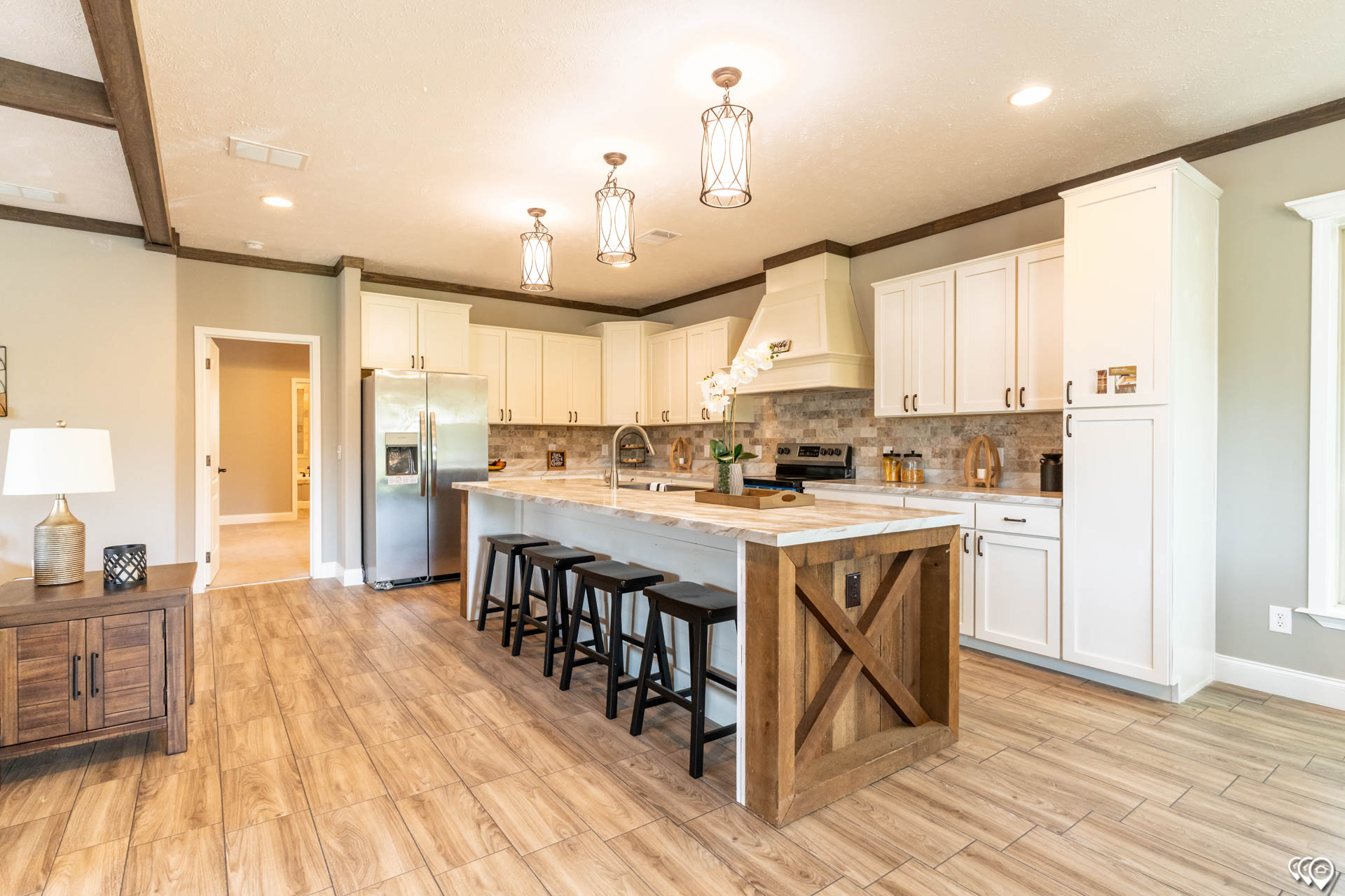 Spacious kitchen with wooden floors, a central island with four stools, and three pendant lights. White cabinets, stainless steel appliances, warm and inviting.