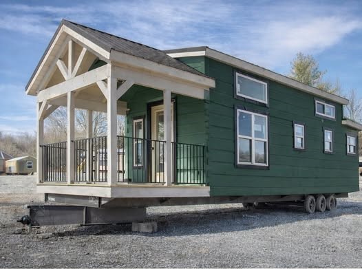 A green tiny house on wheels with a white-trimmed porch and railing. It sits on a gravel lot under a clear sky, conveying a peaceful, mobile lifestyle.
