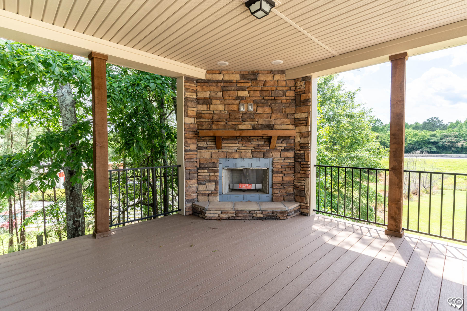 Covered patio with wood decking and a stone fireplace, framed by wooden beams. Overlooks lush trees and a green lawn, conveying a serene atmosphere.
