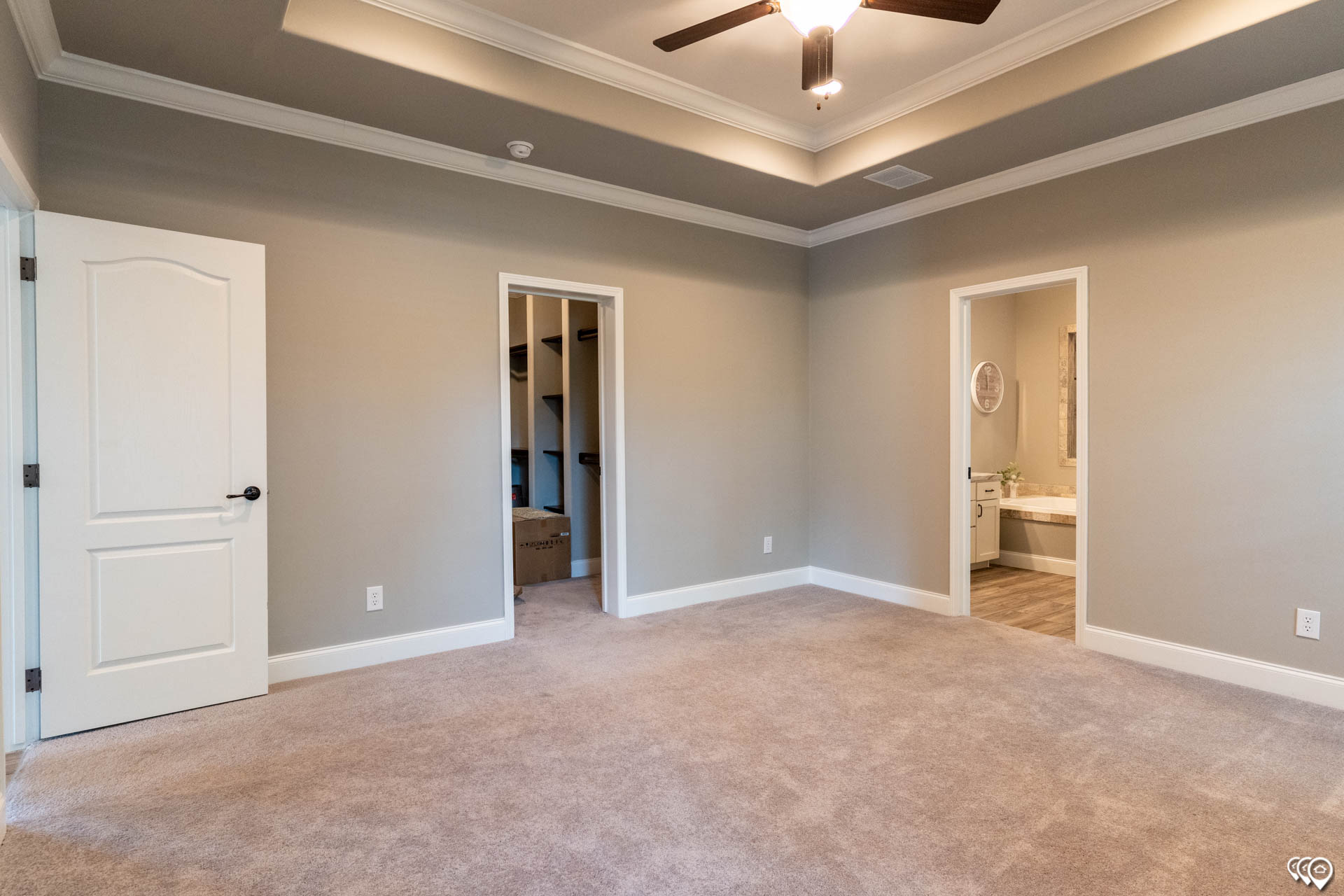 Empty room with beige carpet, gray walls, and a ceiling fan. Two doorways lead to a bathroom and a closet. Warm lighting creates a calm atmosphere.