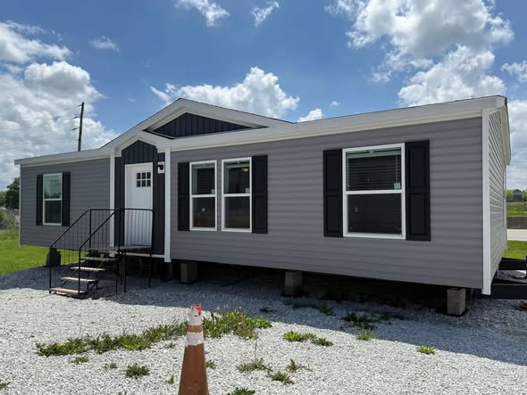 A gray manufactured home with a black roof sits on gravel, featuring black shutters and a white door. Bright day with scattered clouds; calm, inviting scene.