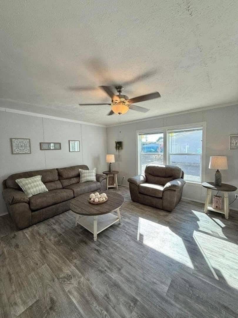 Cozy living room with brown sofas, a wooden coffee table, floor lamps, and a ceiling fan. Sunlight streams through large windows onto the wooden floor.