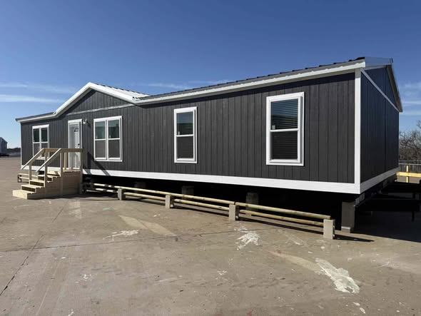 Dark gray modular home with white trim, elevated on supports in a sunny outdoor lot. Features multiple windows and a small wooden staircase.