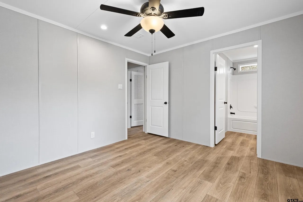 A modern, empty room with light gray walls and wooden flooring. A ceiling fan with a light hangs in the center. Doorways lead to a bathroom and hallway.