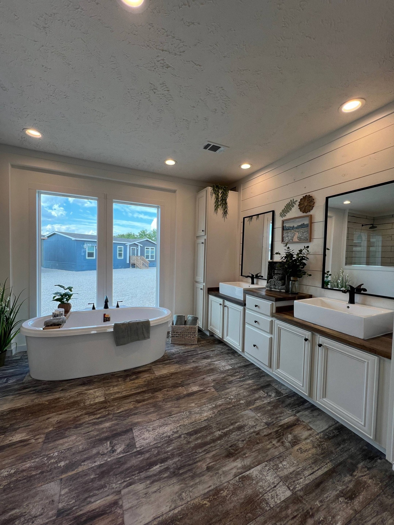Bright, modern bathroom with rustic wood flooring, standalone bathtub, and dual sinks. Large windows reveal an outdoor view, enhancing the airy feel.