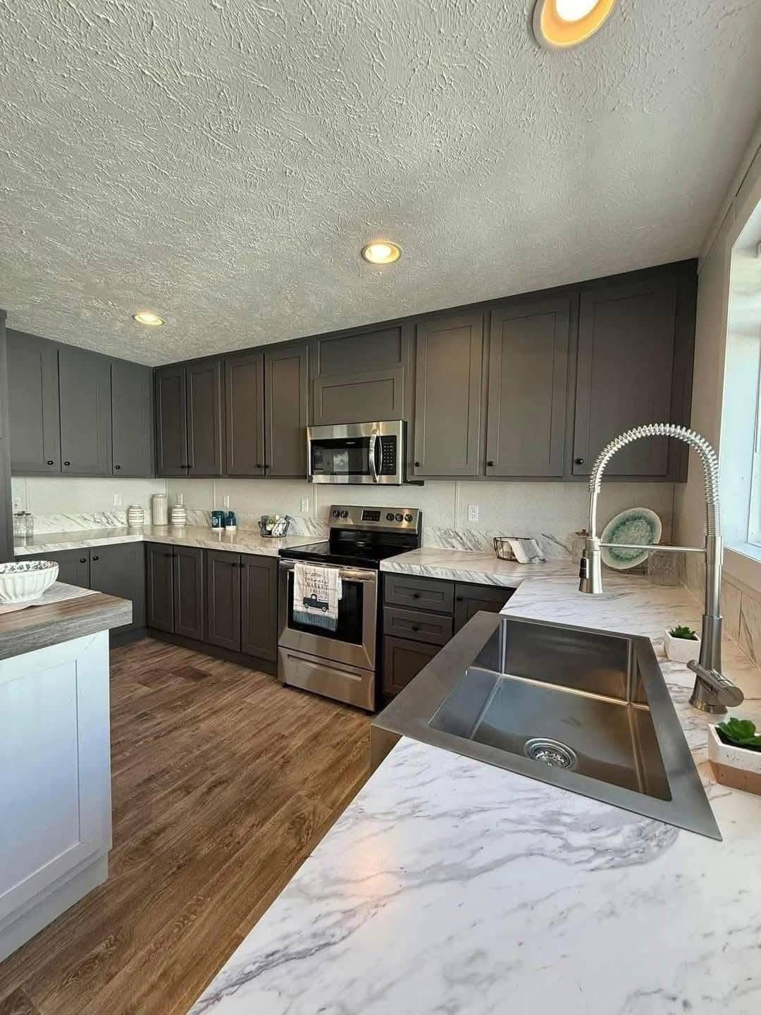 Modern kitchen with dark gray cabinets, marble countertops, and stainless steel appliances. Bright lighting creates a clean, inviting atmosphere.