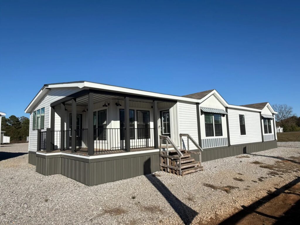 A white manufactured home with a covered porch and dark railings, set on a gravel yard under a clear blue sky, creating a calm, inviting atmosphere.