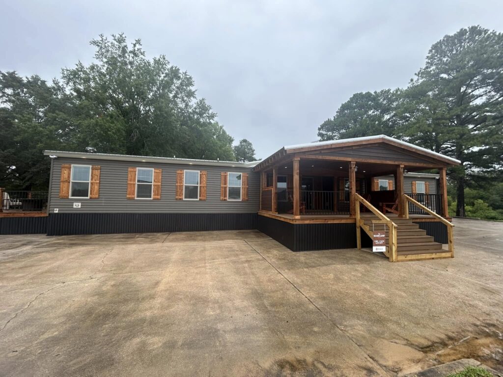 Single-story mobile home with a wide wooden porch and stairs, set against a backdrop of tall trees. The overcast sky creates a calm, serene mood.