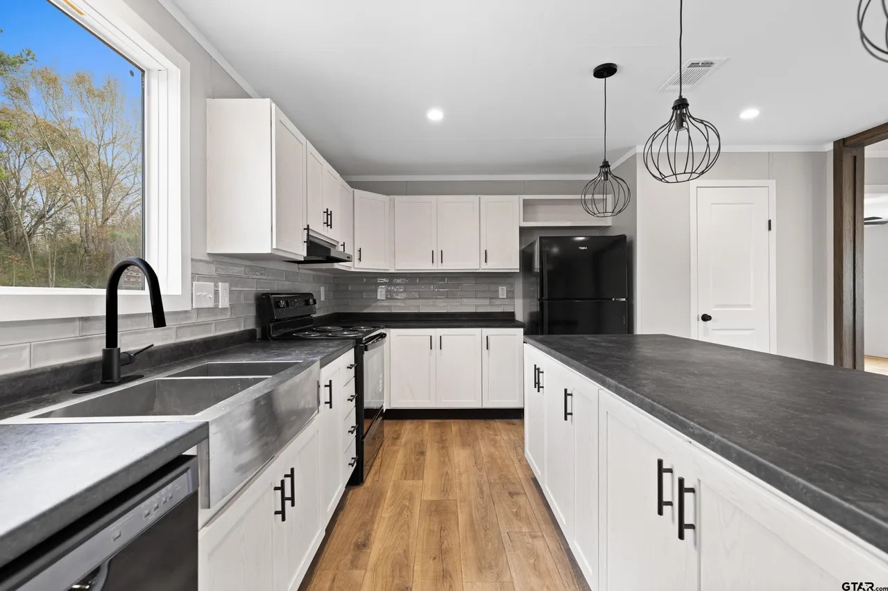 Modern kitchen with white cabinets, black countertops, and wooden floors. Features a large island, pendant lights, and a window with trees outside.