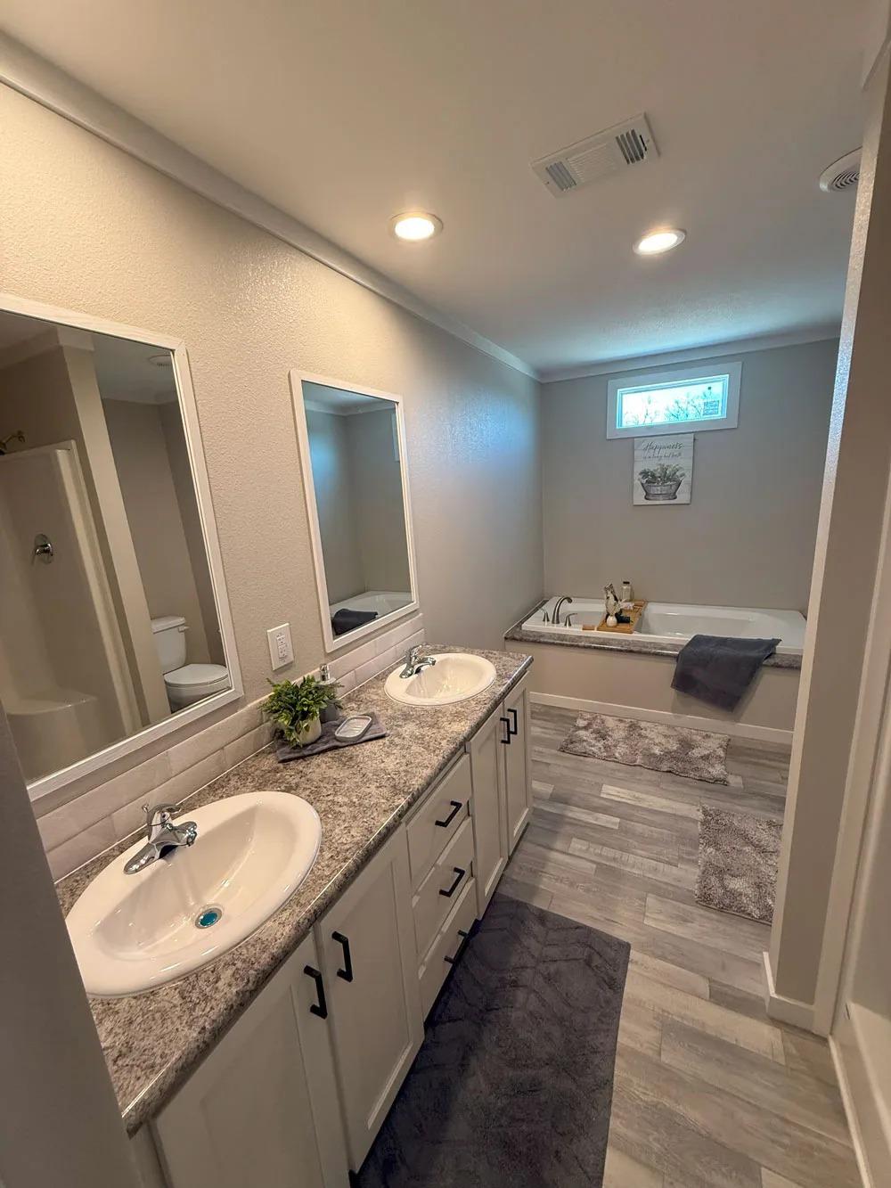 Modern bathroom featuring a granite double-sink vanity with mirrors. A sleek bathtub is set against a window, surrounded by soft gray mats, creating a calming ambiance.