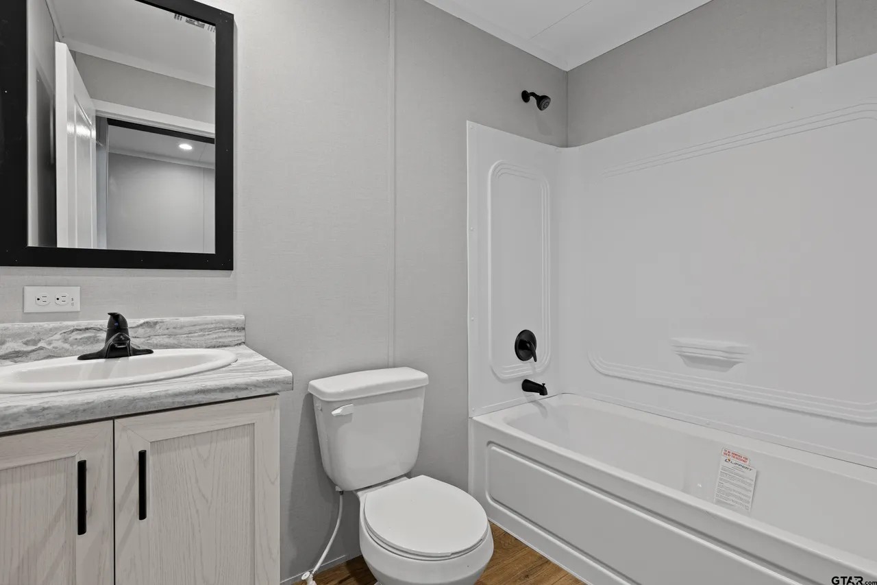 Modern bathroom with soft gray walls. Features include a white bathtub with black fixtures, a toilet, and a sink with a gray countertop and black-framed mirror. The tone is clean and minimalist.