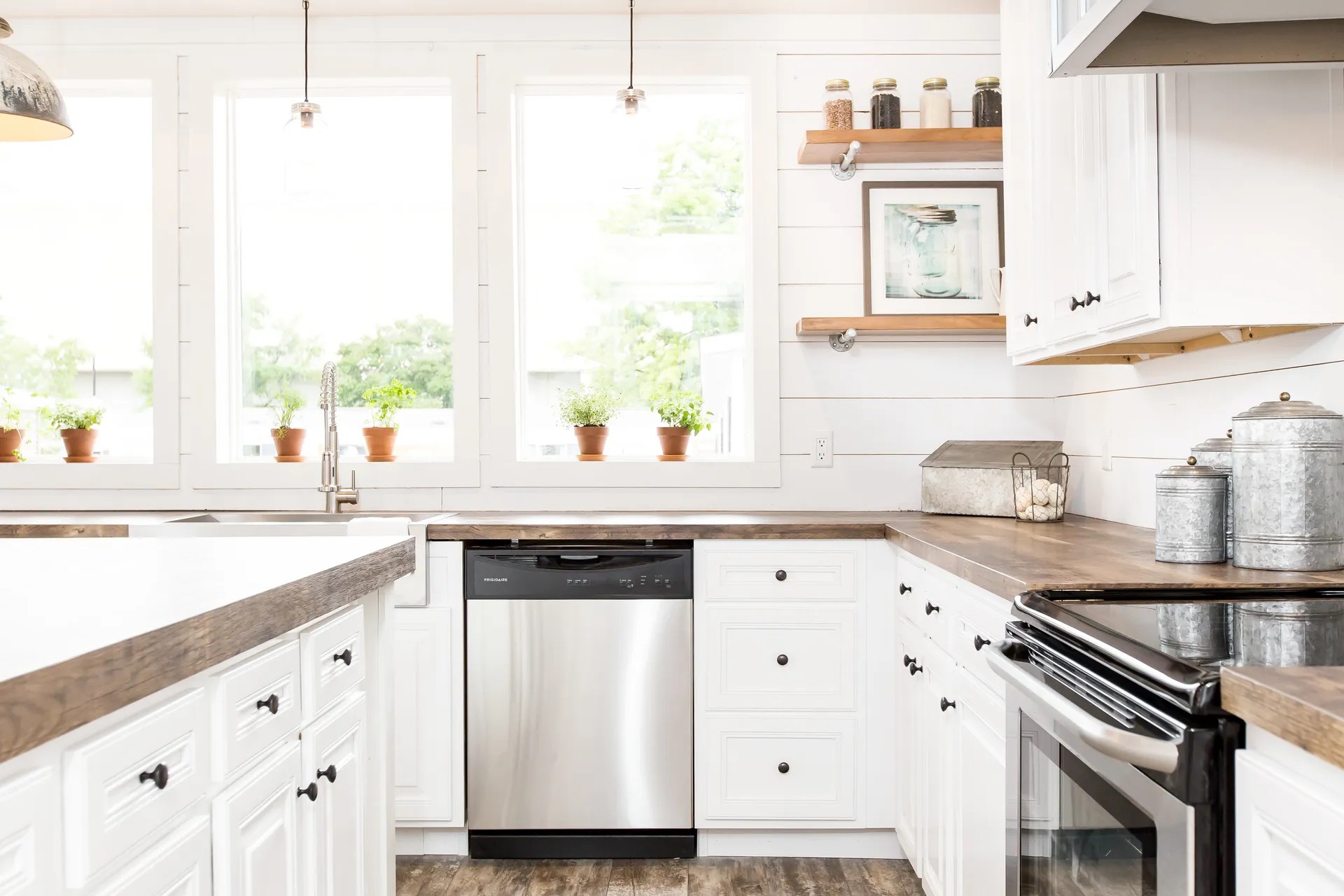 Bright farmhouse kitchen with white cabinets, wood countertops, and stainless steel appliances. Potted plants line the window sill, adding a fresh touch.
