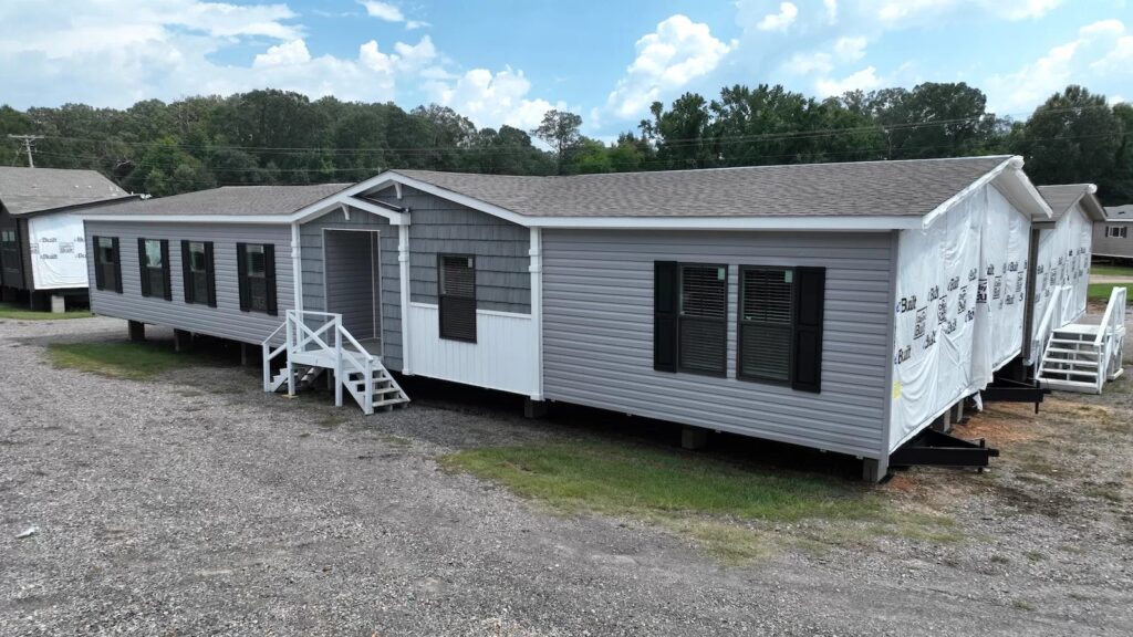 A gray manufactured home with white trim and dark shutters, positioned on a gravel lot. Stairs lead to the entry. Trees and a blue sky are in the background.