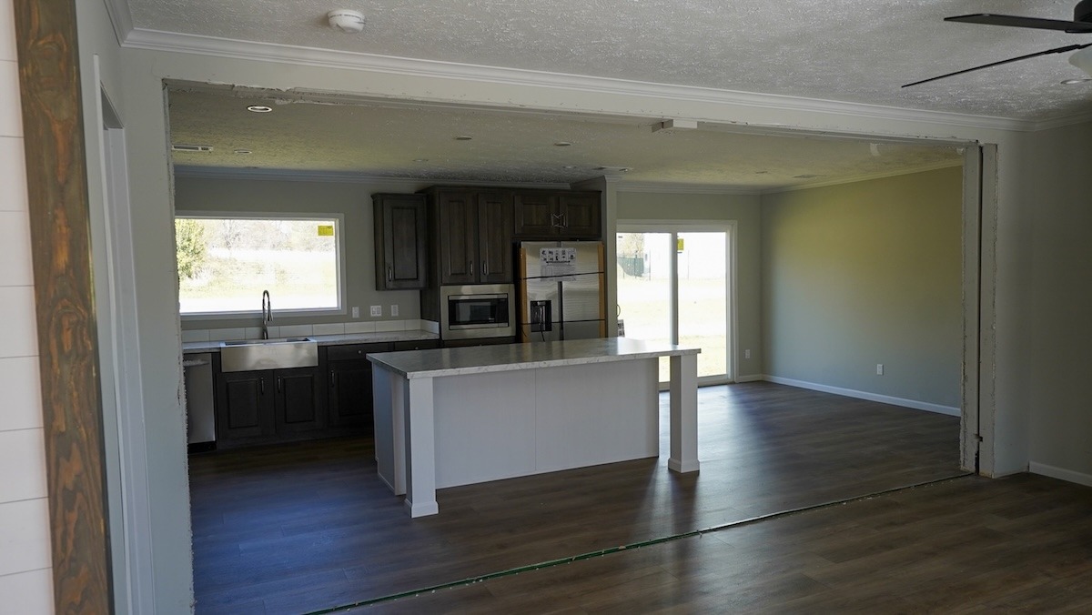 Open, modern kitchen and living area with light wood flooring. Features a central island, dark cabinets, large windows, and a ceiling fan, conveying a spacious, airy feel.