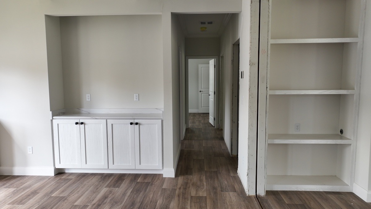 Empty room with light wood flooring and neutral walls. Features white built-in cabinets and shelves. Leads to a hallway with several doors. Soft lighting creates a calm atmosphere.