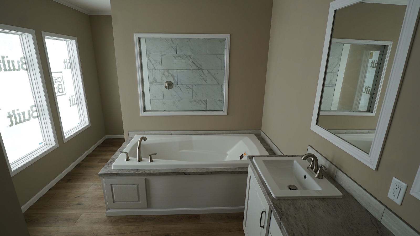 A modern bathroom with a bathtub featuring a marble surround, adjacent to a white sink with a silver faucet and mirror. Two frosted windows provide soft light.