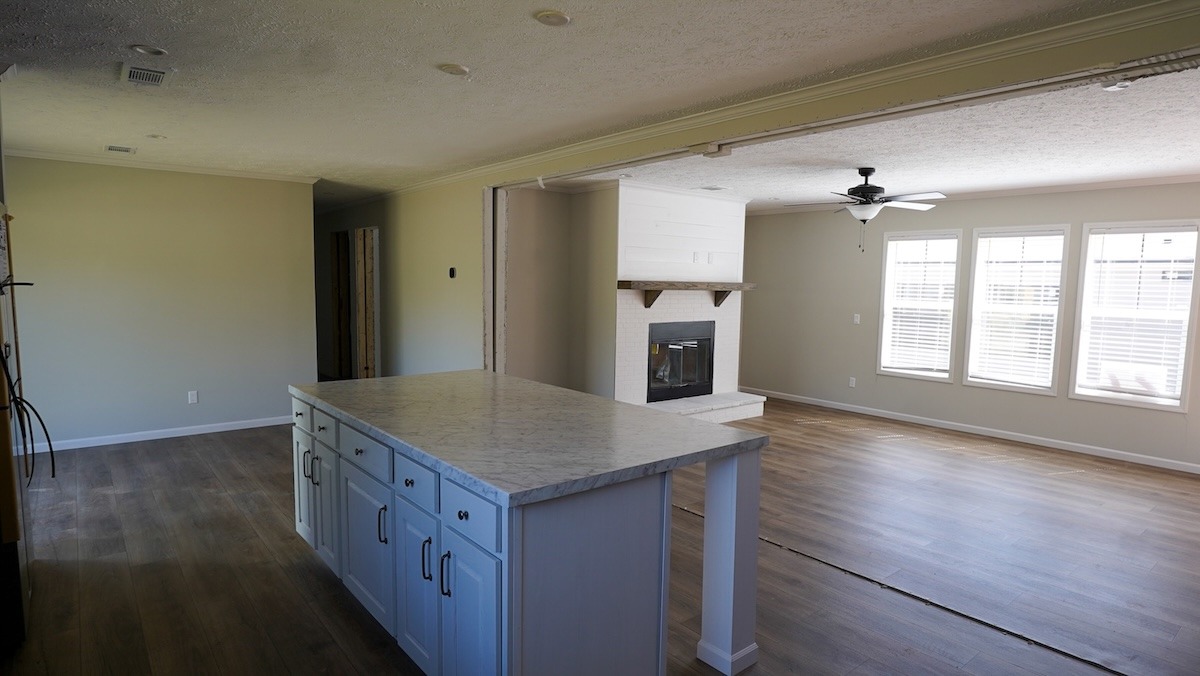 Spacious, sunlit open floor plan with a kitchen island, hardwood floors, and a cozy fireplace surrounded by large windows, creating a warm ambiance.