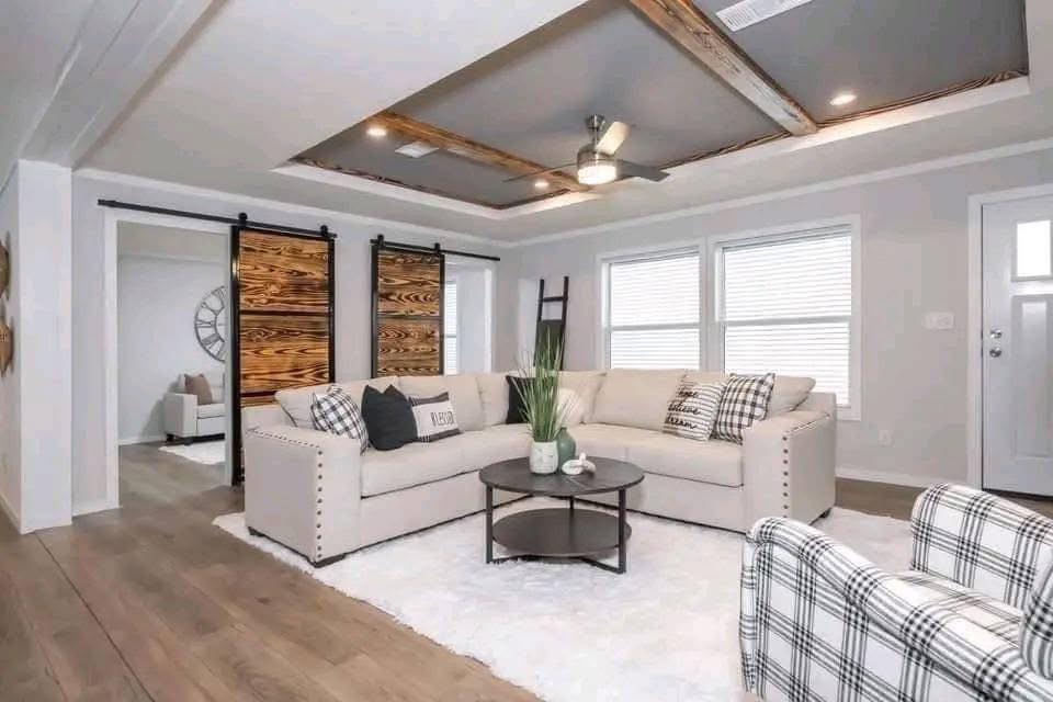Modern living room with a beige sectional sofa, plaid armchair, and round coffee table on a white rug. Barn doors and tray ceiling add rustic charm.