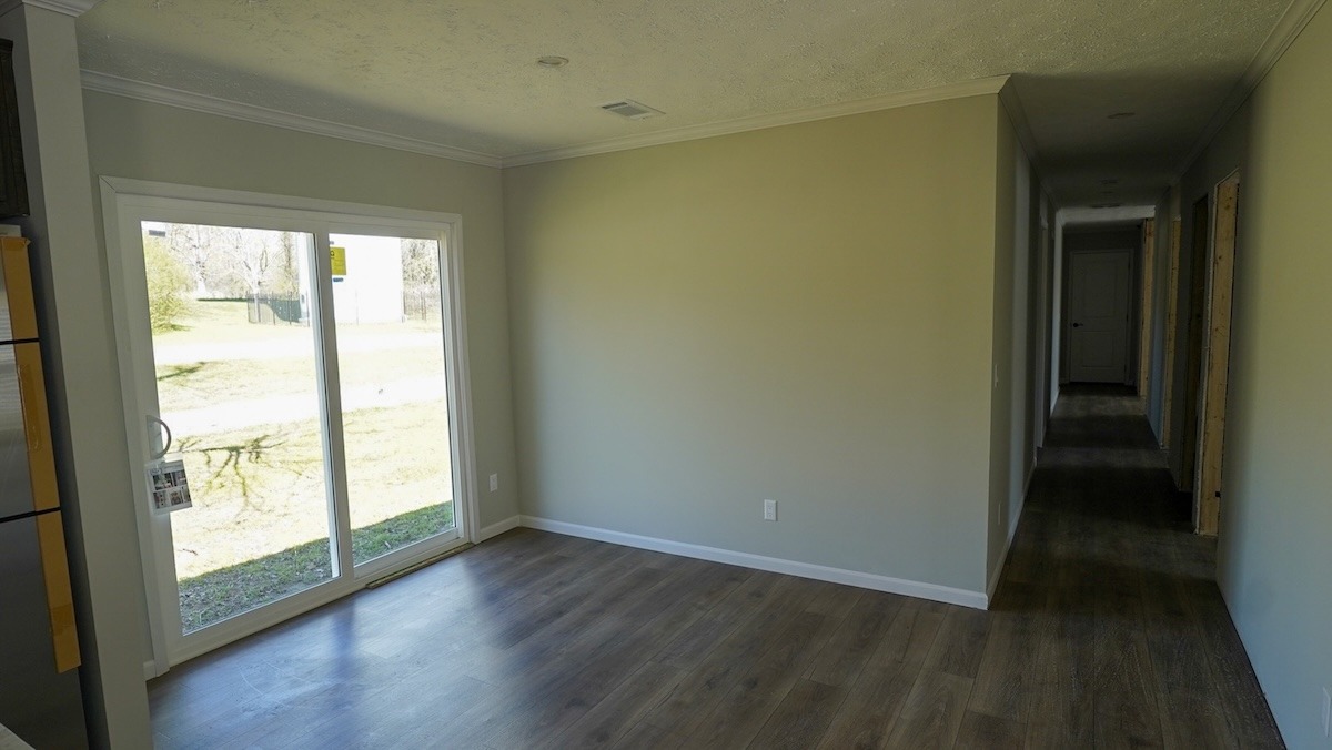 A bright, empty room with large sliding glass doors on the left, leading outside, and a long hallway to the right. The wooden floor adds warmth.