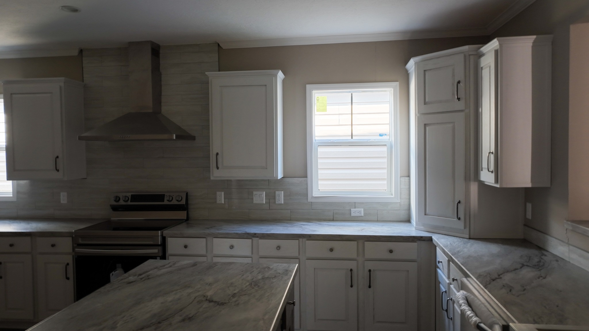 Bright kitchen featuring white cabinets, gray marble countertops, and stainless steel stove with a vent hood. Sunlight streams through two windows.