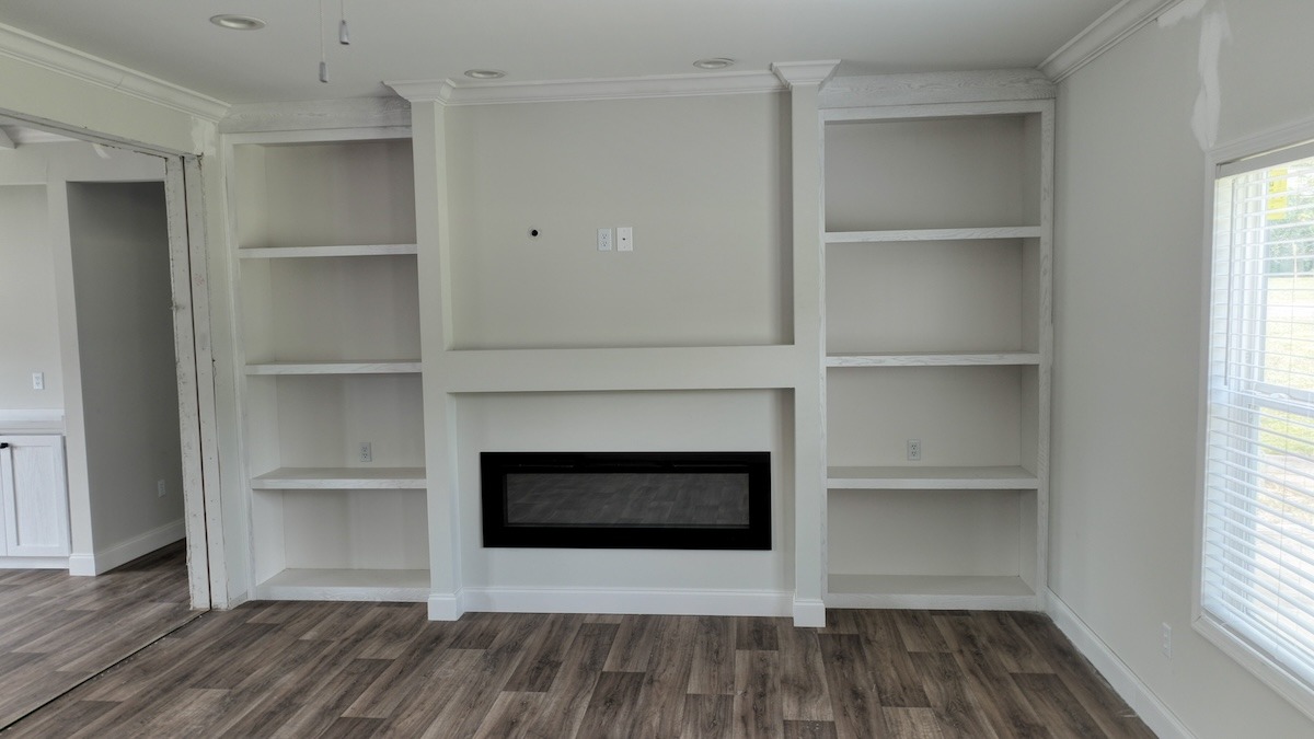 A modern living room features white built-in shelves surrounding a sleek, recessed fireplace. The space is brightened by natural light from nearby windows.