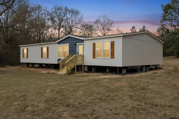 A modern prefab home with white siding and wooden shutters sits on a grassy field. A small wooden porch leads to the front door. The sky is a colorful sunset.