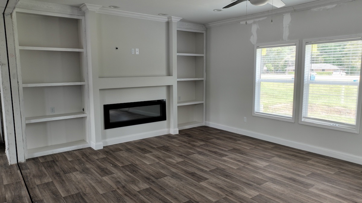 Spacious living room with light gray walls, built-in shelves, and a sleek electric fireplace. Large windows bring in natural light. Cozy and modern ambiance.