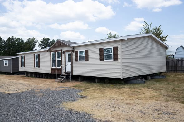 A single-story beige mobile home with dark brown shutters and a small porch is set on a gravel and dirt lot. Overcast sky and surrounding trees add a serene tone.