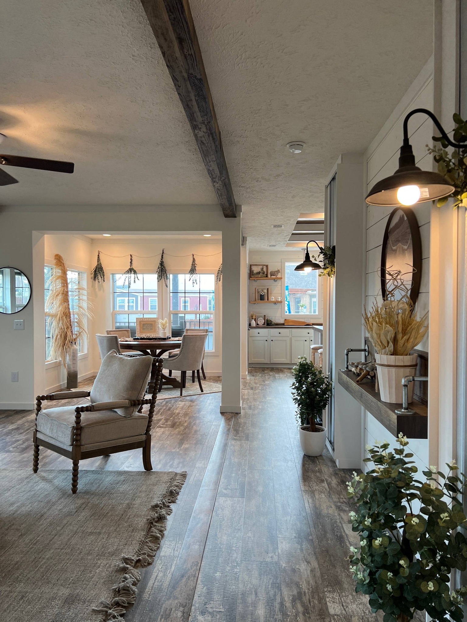A cozy living space with rustic wooden flooring and beams. The area features elegant chairs, a dining table by large windows, and kitchen with white cabinets. The light, airy atmosphere is enhanced by plants and warm lighting.