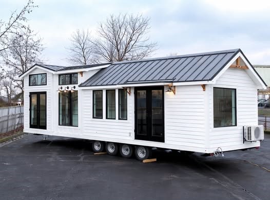 A modern tiny house on wheels with a white exterior and black trim, featuring multiple windows and a metal roof, parked on an asphalt surface.