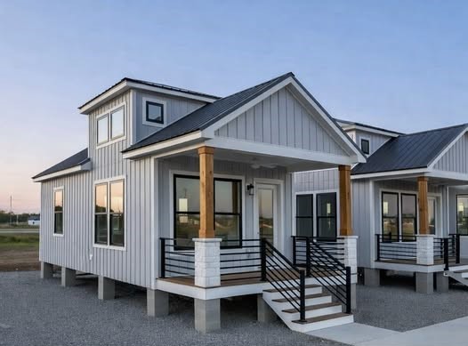 Modern tiny homes on stilts with gray siding and black metal roofs. Each has a small porch with wooden columns and a ramp. The sky is clear, suggesting a peaceful setting.