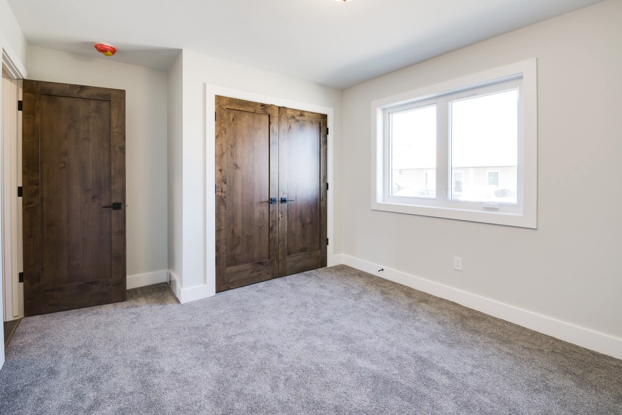 Small empty room with gray carpet, two wooden closet doors, and a wooden interior door. A large window lets in natural light, creating a bright, minimalist space.