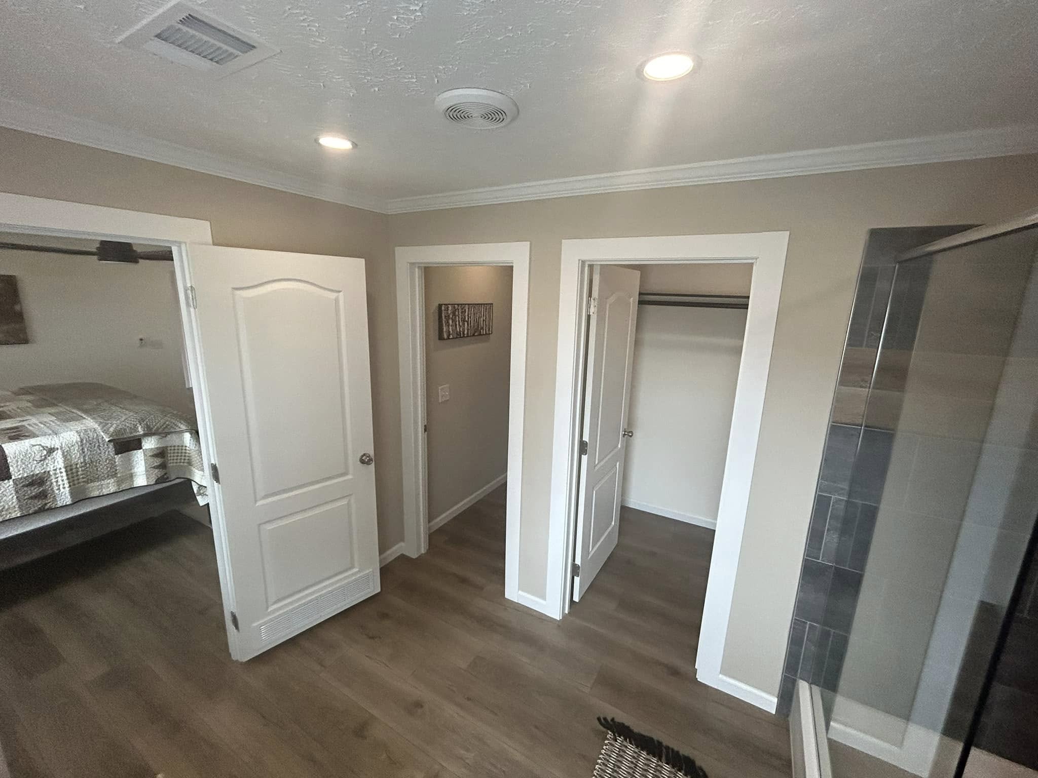 A cozy room with light wood flooring and beige walls. Open doors reveal a bedroom with a quilted bed and an empty closet. Soft, warm lighting creates a welcoming atmosphere.