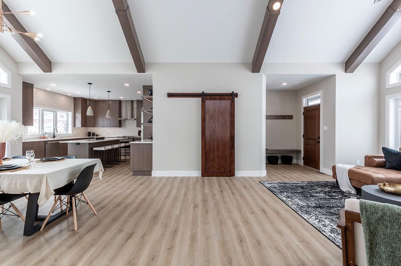 Open-concept living space with light wood floors, exposed beams, and a sliding barn door. Modern kitchen, dining table, and cozy seating area. Bright and inviting.