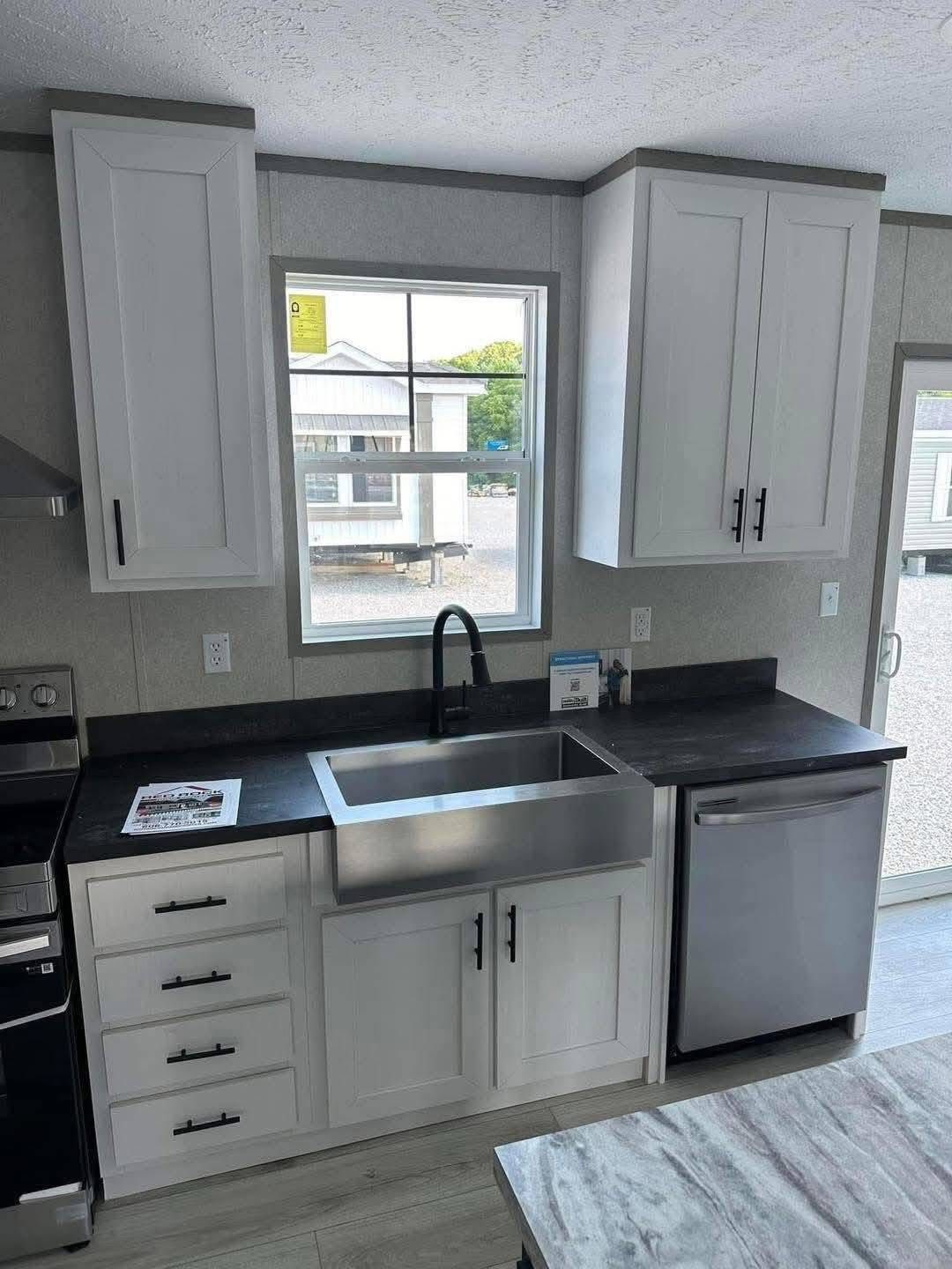 Modern kitchen with white cabinets, a stainless steel farmhouse sink, dark countertops, and a built-in dishwasher. A window provides natural light.