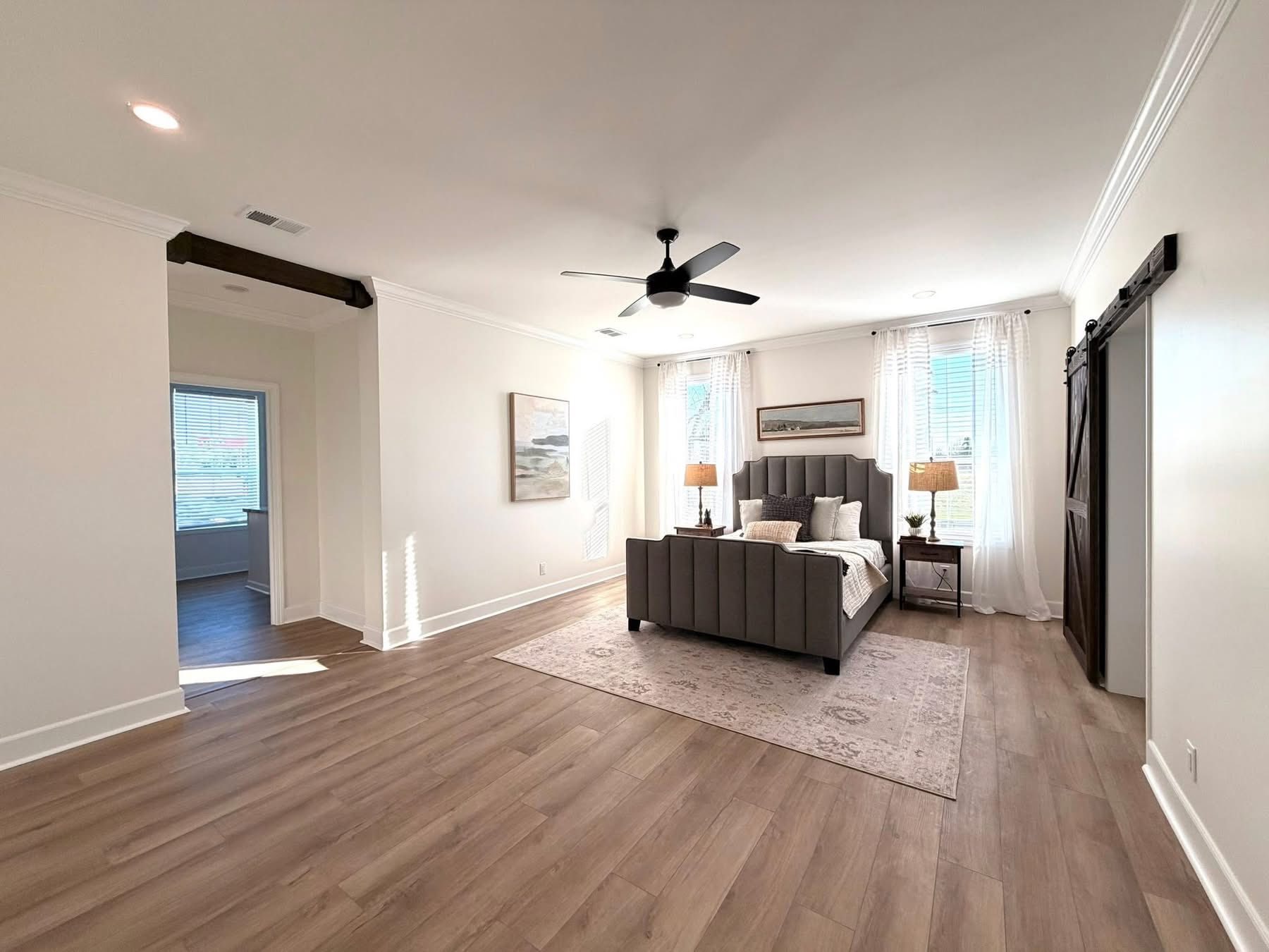 Spacious bedroom with gray bed, pillows, and side tables with lamps. Wooden floor, ceiling fan, large windows with sheer curtains, and soft lighting. Cozy and inviting atmosphere.