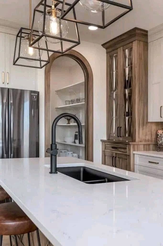 Modern kitchen with white marble island, black faucet, and geometric pendant lights. Wooden cabinets and stainless steel fridge add warmth and style.