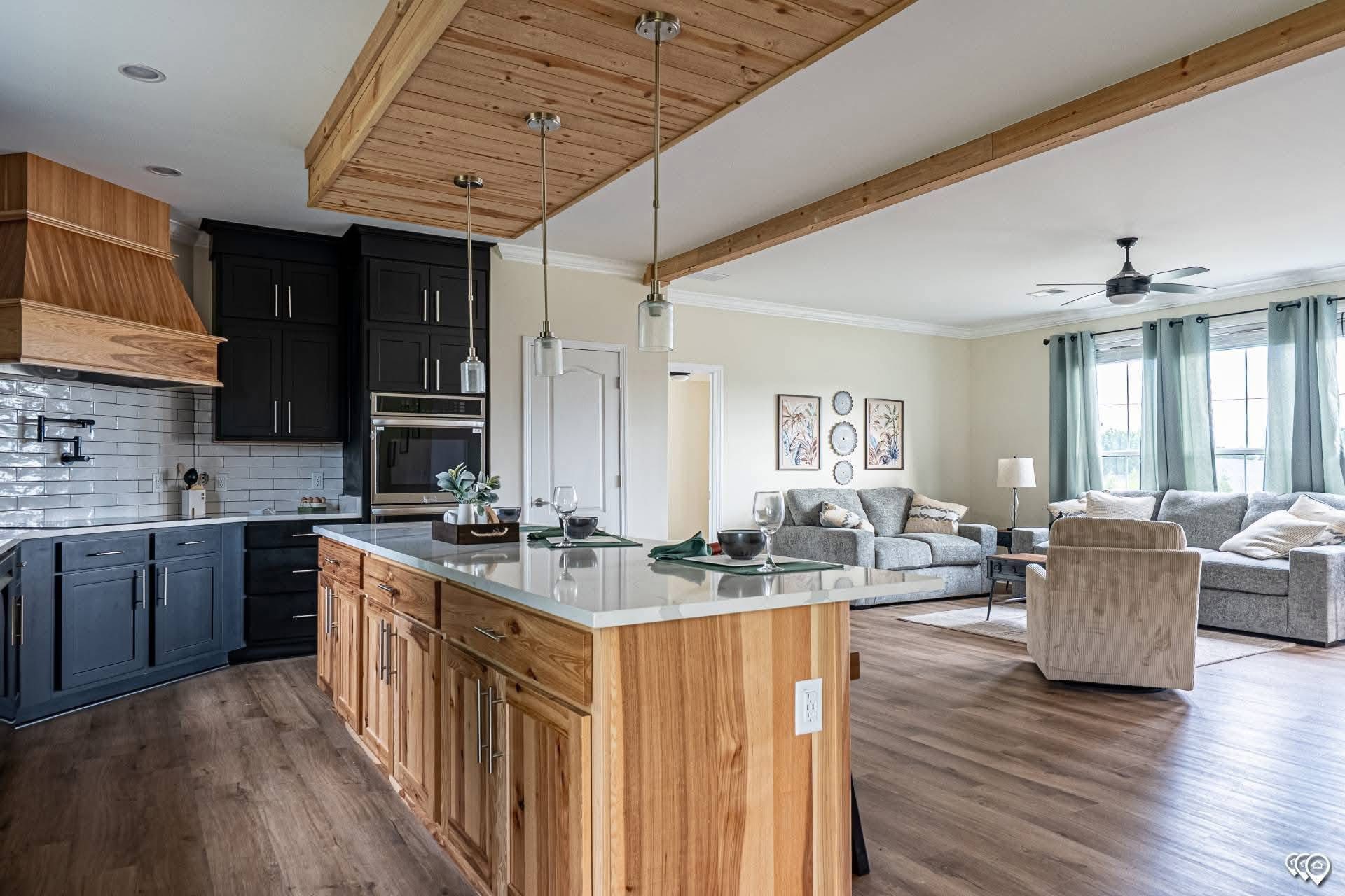 Spacious open-concept kitchen and living room with wooden flooring. The kitchen features dark cabinets and a light wood island. The living room has gray sofas, green curtains, and wall art, creating a cozy, modern feel.