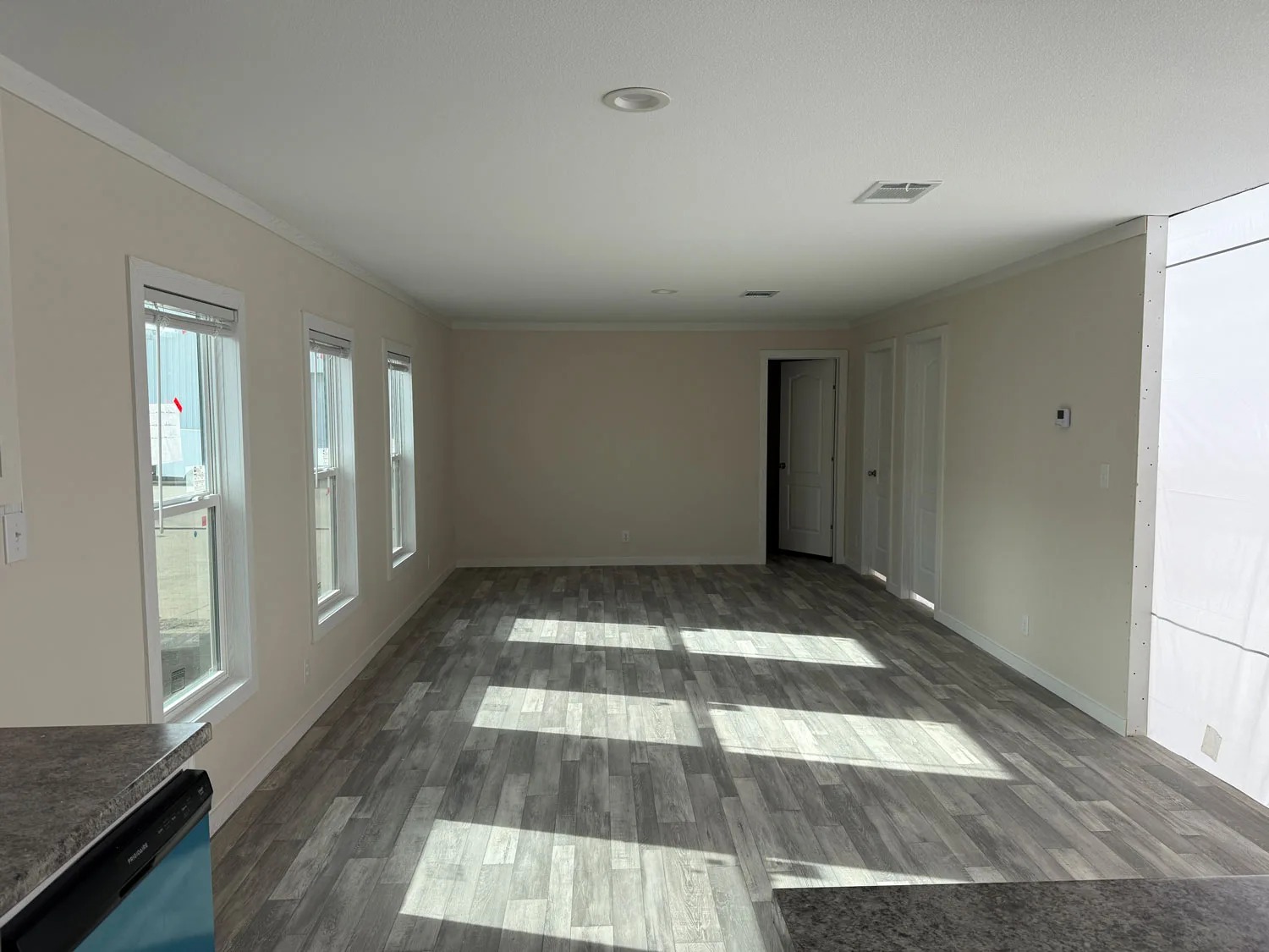 Empty room with four windows on the left wall casting sunlight onto a gray wooden floor. Neutral walls and multiple closed doors can be seen. Bright and airy ambiance.