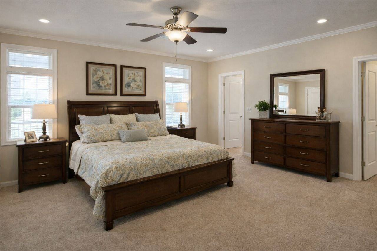 A cozy bedroom with a wooden bed, matching dresser, and nightstands. Beige carpet, floral decor, a ceiling fan, and soft lighting create a warm ambiance.