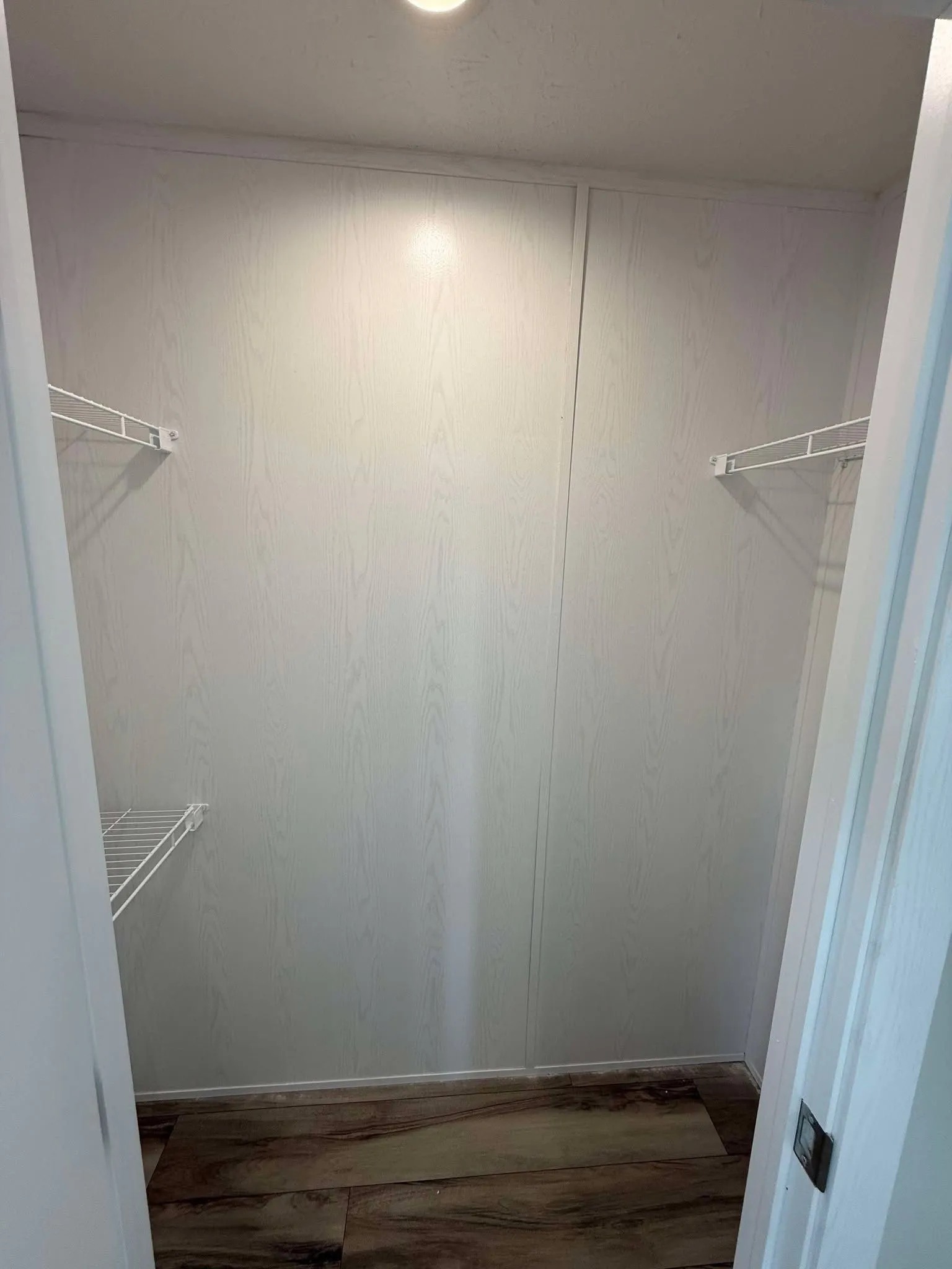 Empty white closet with wood-patterned floor, lit by ceiling light. Two metal wire shelves on left and right walls, creating a spacious, minimalist feel.