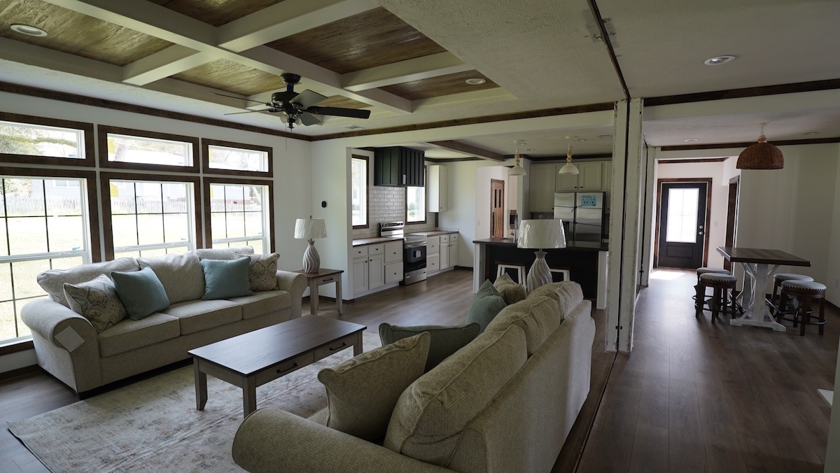 Stylish open-plan living room with beige sofas, light blue cushions, wooden coffee table. Bright kitchen with black accents, bar stools, and wood flooring. Cozy and inviting.