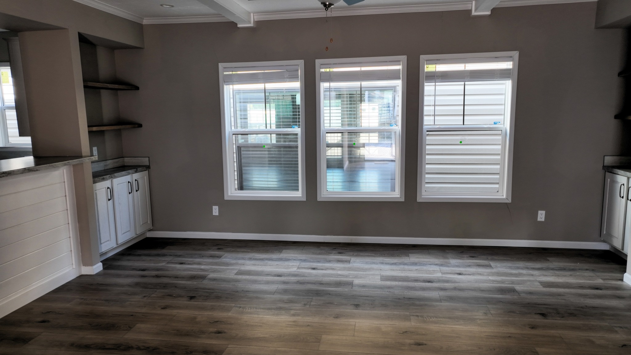 A spacious room with dark wood flooring, three large windows with white blinds, beige walls, and white trim. Built-in shelves and cabinets add storage.