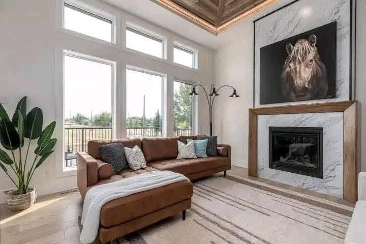 A modern living room with a brown leather sectional sofa, a floor lamp, and cushions. Large windows brighten the space with natural light. A plant is by the window, and a marble fireplace with a horse painting above adds elegance.