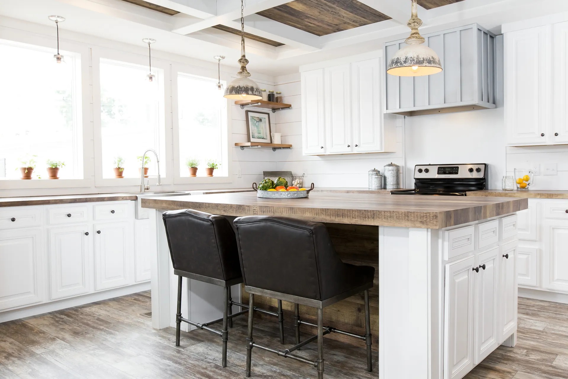 Bright, rustic kitchen with white cabinets, wood countertops, and an island with two black chairs. Hanging lights add warmth; potted plants line windows.