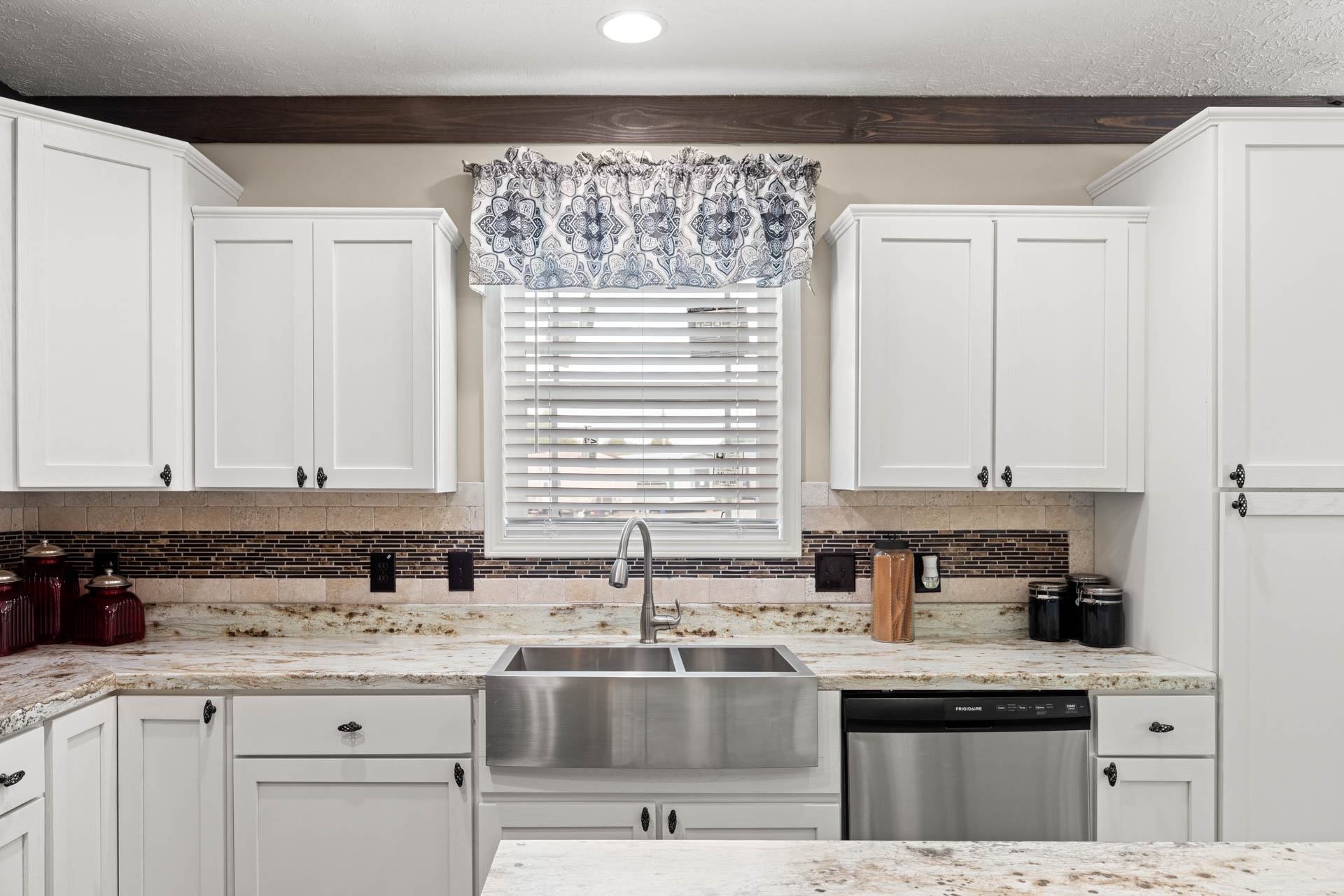 Bright kitchen with white cabinets and a marble countertop. Central window with patterned valance, stainless steel sink, and a modern, clean ambiance.